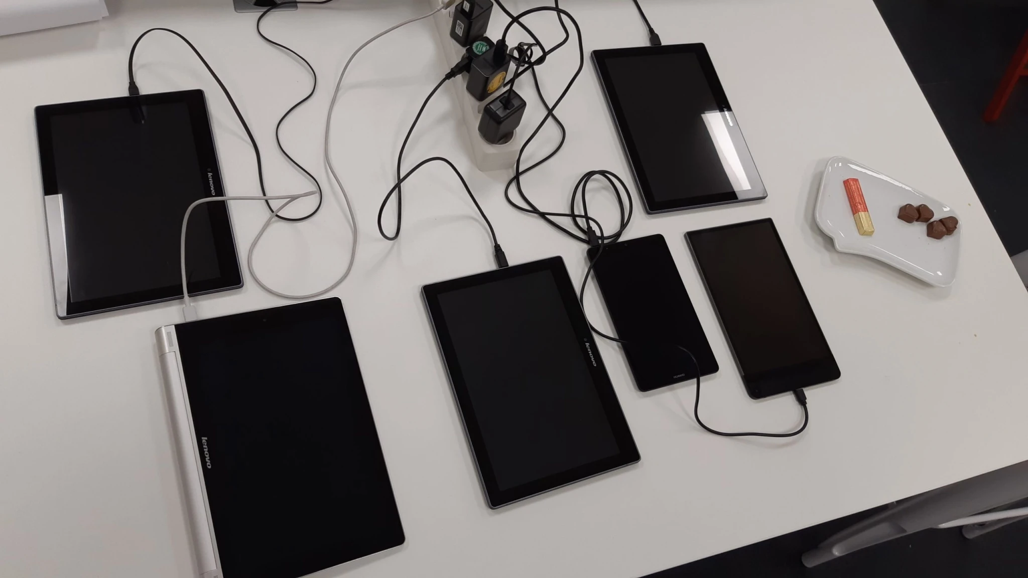 Mehrere Tablets und Smartphones auf einem Tisch, verbunden mit Ladekabeln – Vorbereitung auf Tests.