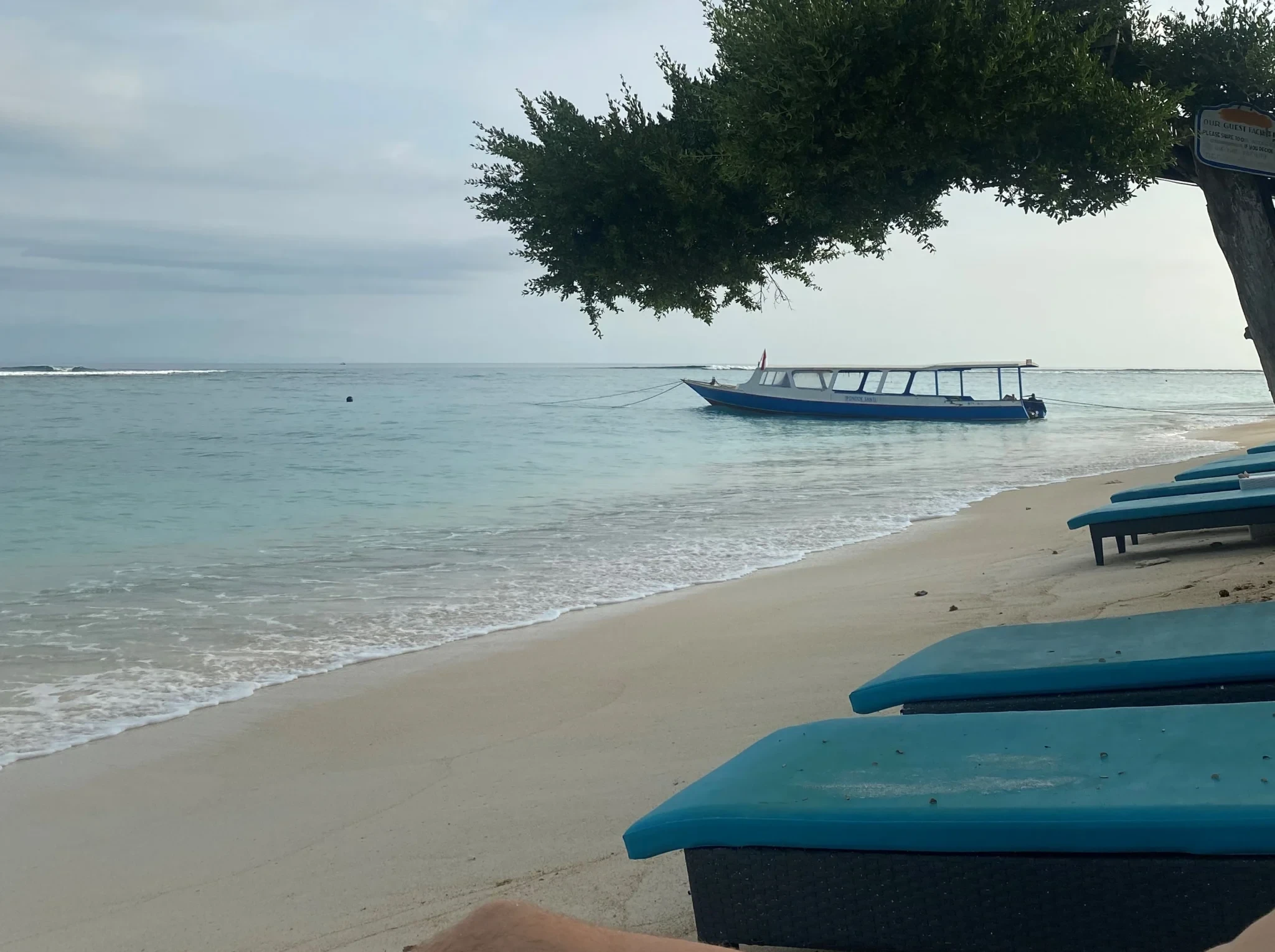Leere Sonnenliegen am Strand von Bali mit Blick aufs ruhige Meer und ein traditionelles Boot bei bewölktem Himmel.